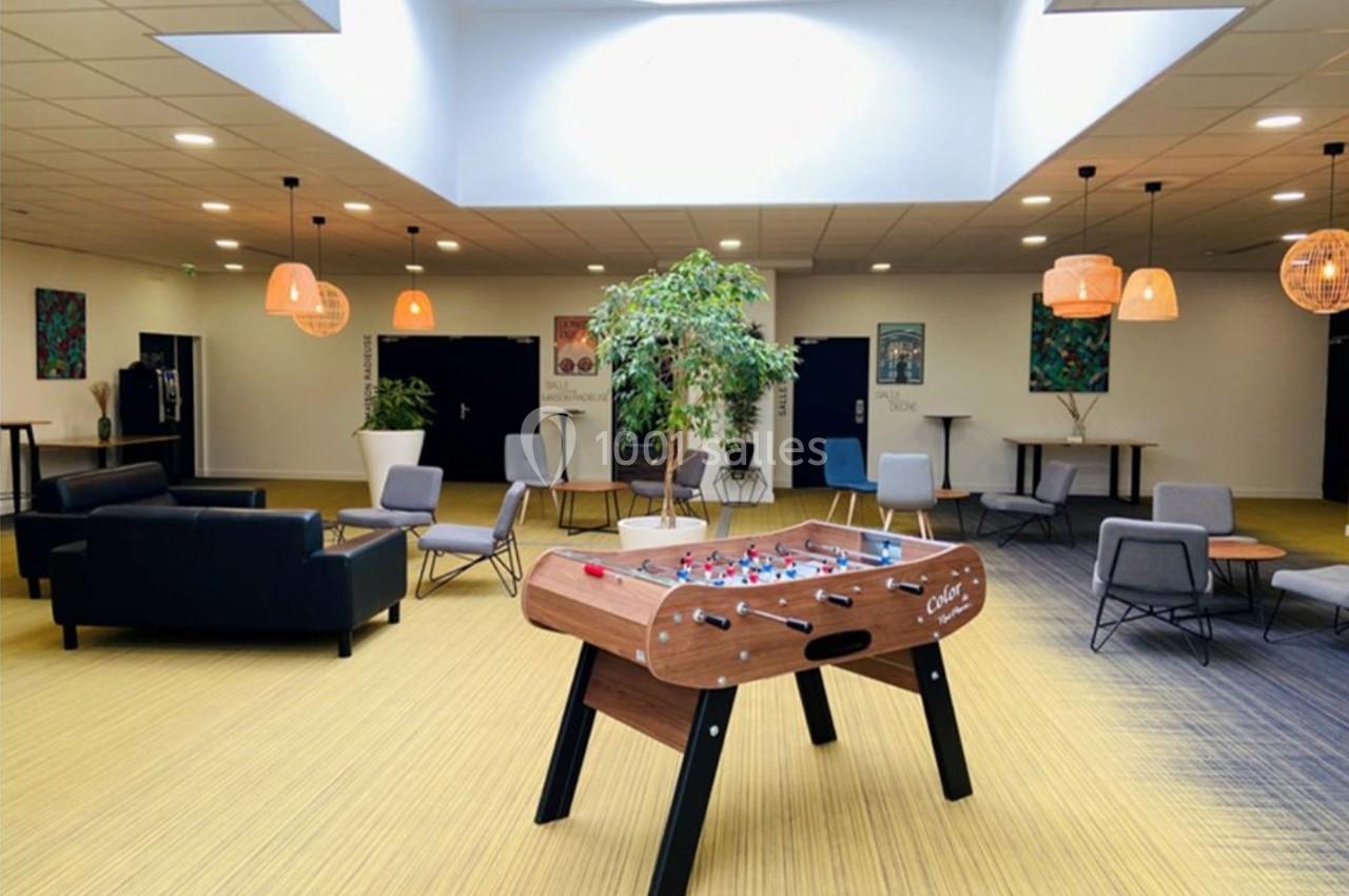 Salle lumineuse avec baby-foot au centre, entourée de fauteuils, tables et plantes sous un puits de lumière.