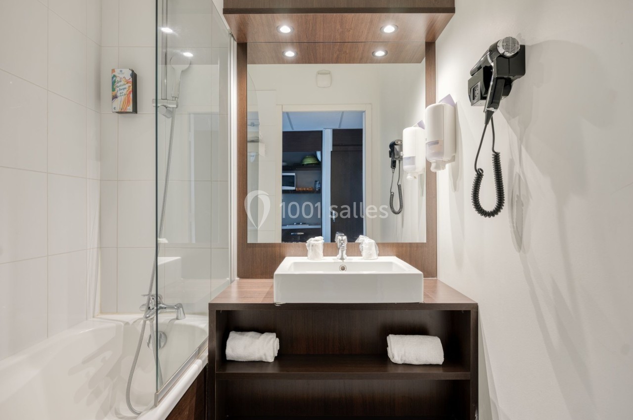 Salle de bain moderne avec baignoire, lavabo sur meuble en bois, miroir éclairé et sèche-cheveux mural.