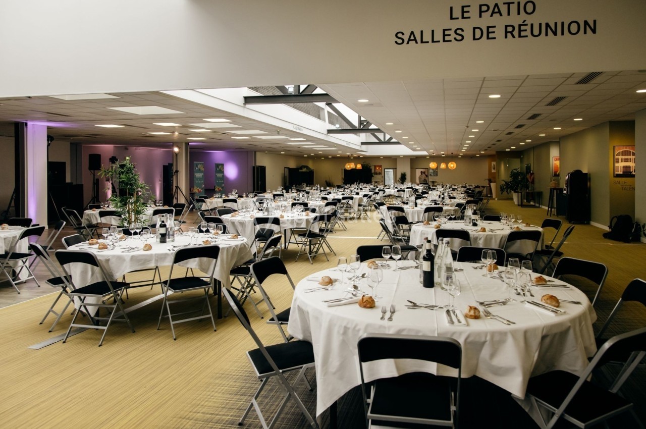 Salle de réception lumineuse avec tables rondes dressées pour un événement, chaises noires et décoration sobre.