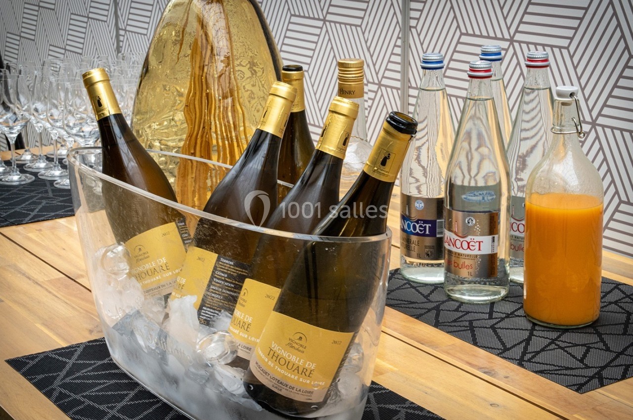 Bouteilles de vin blanc dans un seau à glace, entourées de bouteilles d'eau et d'un pichet de jus d'orange sur une table.