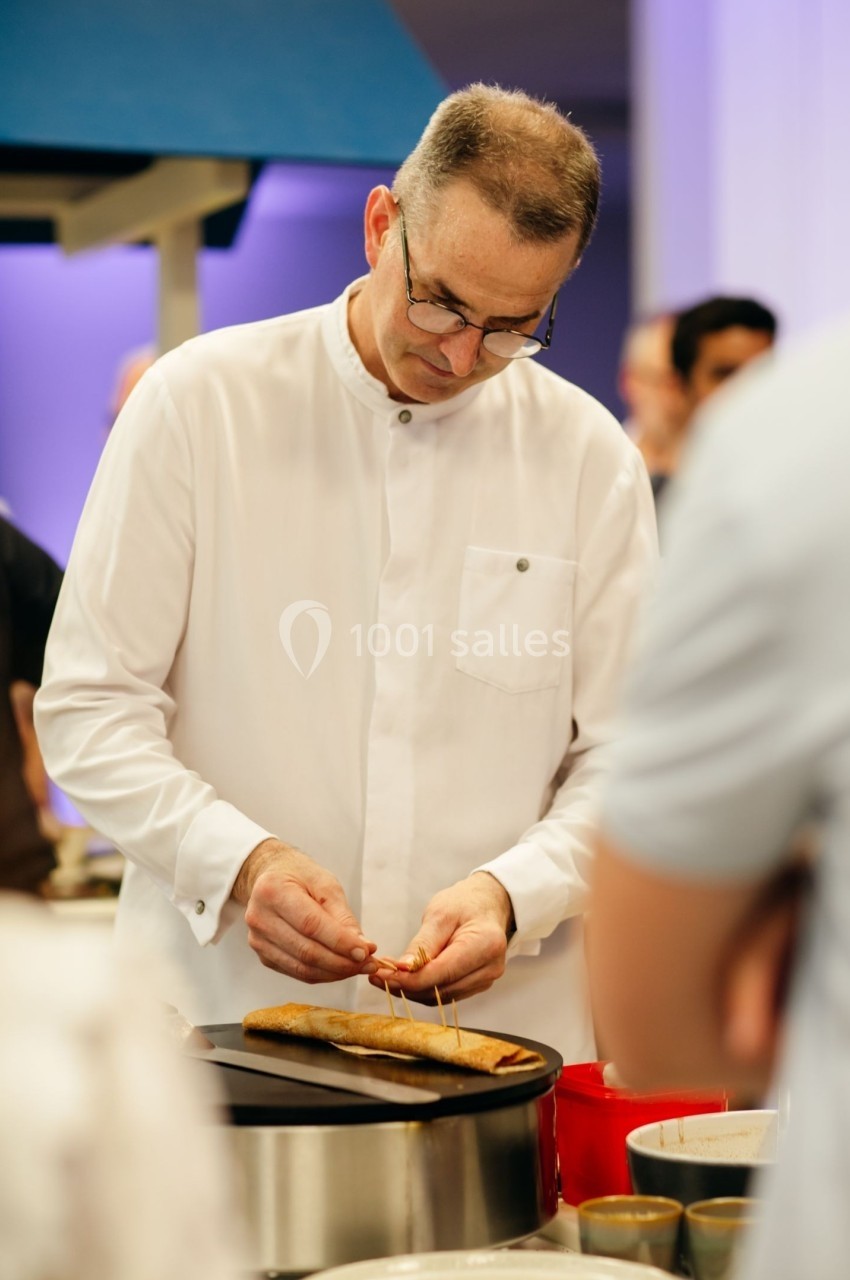 Un chef en veste blanche prépare une crêpe sur une plaque chauffante dans un environnement lumineux.