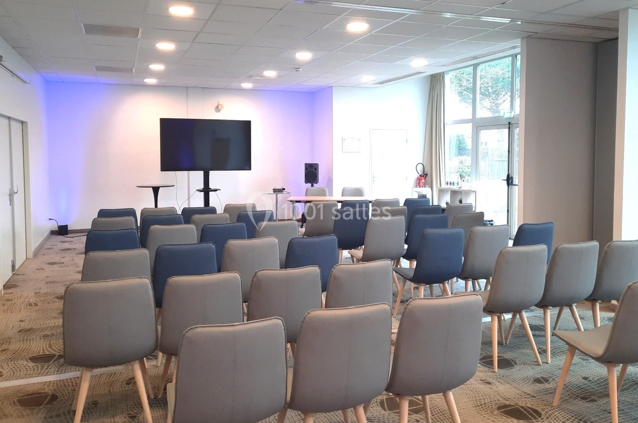 Salle de conférence lumineuse avec des rangées de chaises, un écran et un pupitre devant un mur éclairé en bleu.