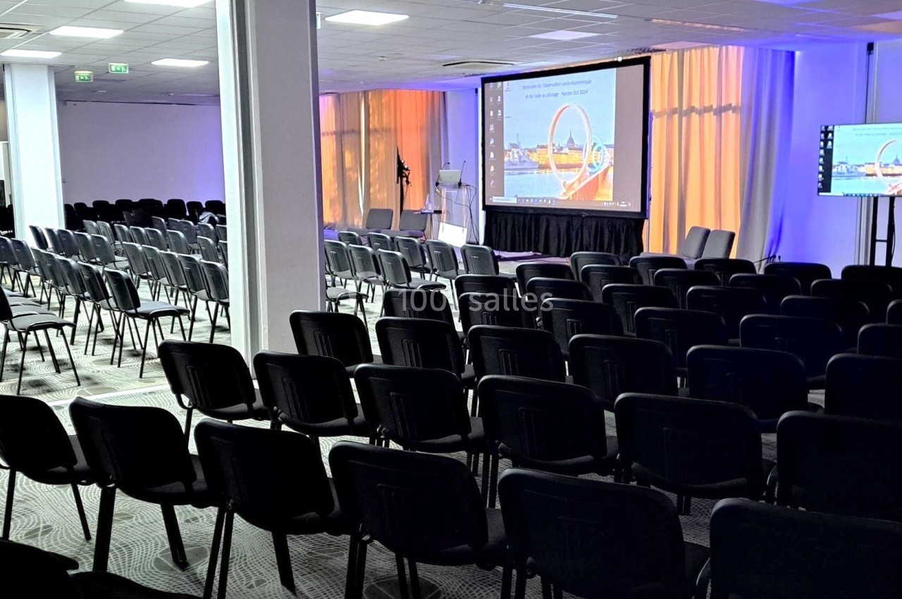 Salle de conférence vide avec des rangées de chaises noires, un écran de projection et un écran secondaire allumés.