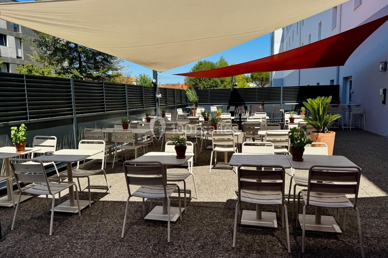 Terrasse extérieure avec tables et chaises blanches, ombragée par des voiles tendues, entourée de plantes et de panneaux.