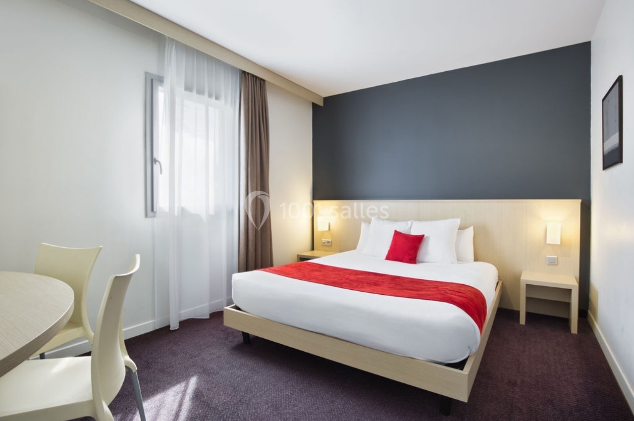Chambre d'hôtel lumineuse avec un lit double, draps blancs, plaid rouge, murs clairs et un mur d'accent gris foncé.