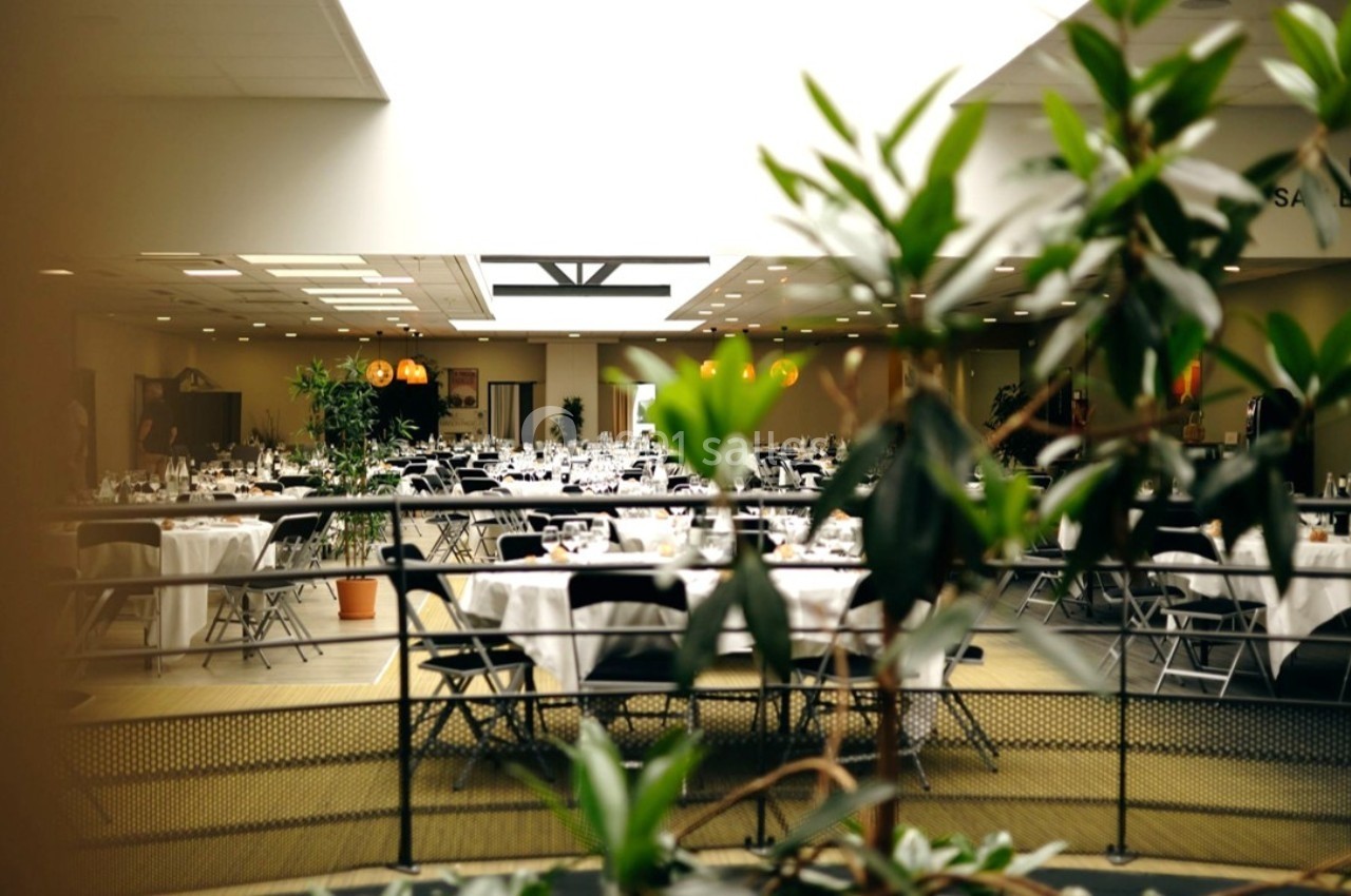 Salle de réception lumineuse avec tables rondes dressées, chaises noires et plantes en premier plan.