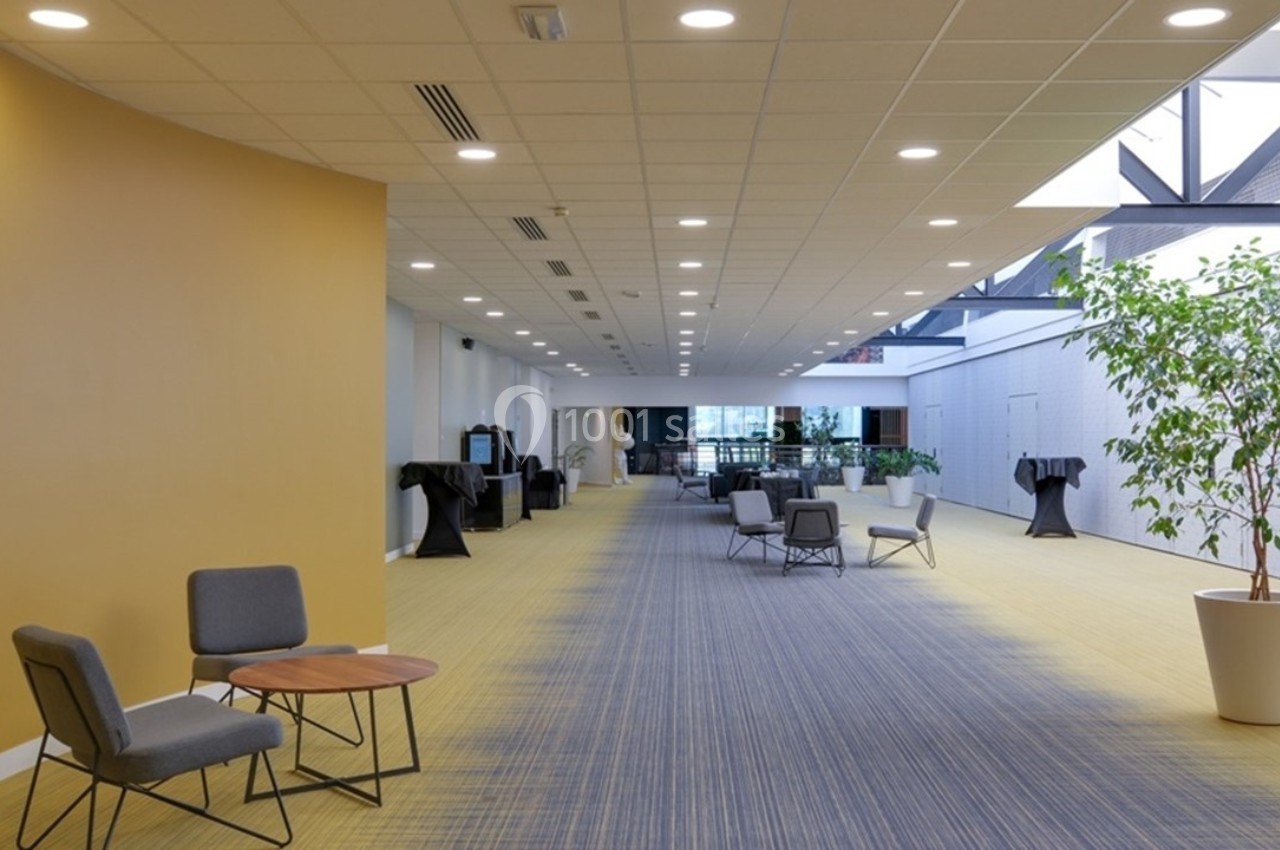 Couloir spacieux avec sol en moquette, chaises et tables basses, éclairage au plafond et plantes en pots.