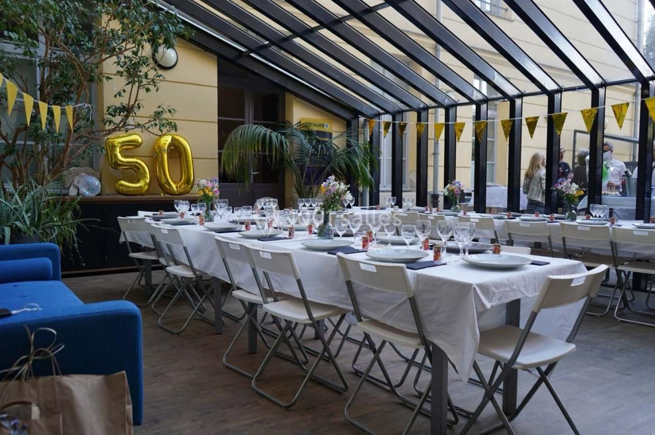 Salle décorée pour une fête avec des tables dressées, des ballons dorés ’50’ et des guirlandes jaunes.