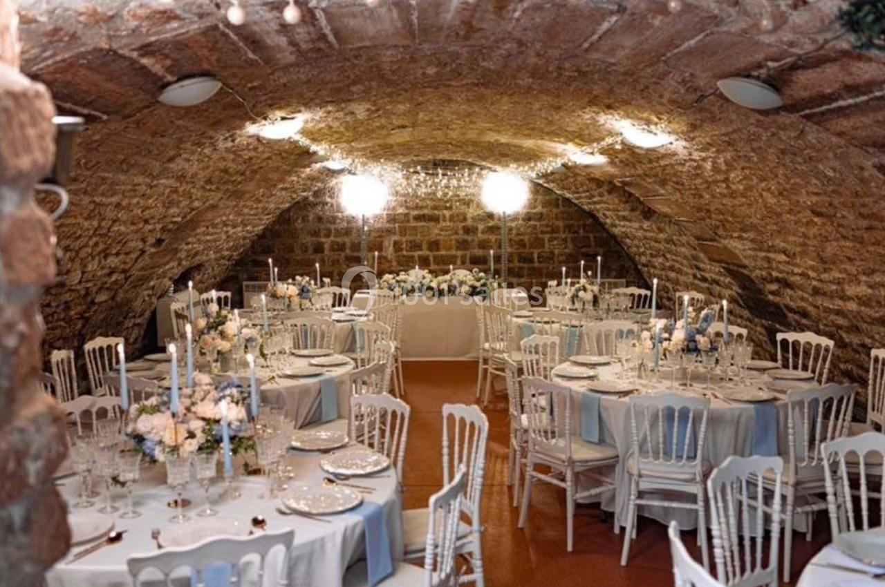 Salle voûtée décorée pour un mariage, avec tables rondes dressées, nappes bleues et arrangements floraux élégants.