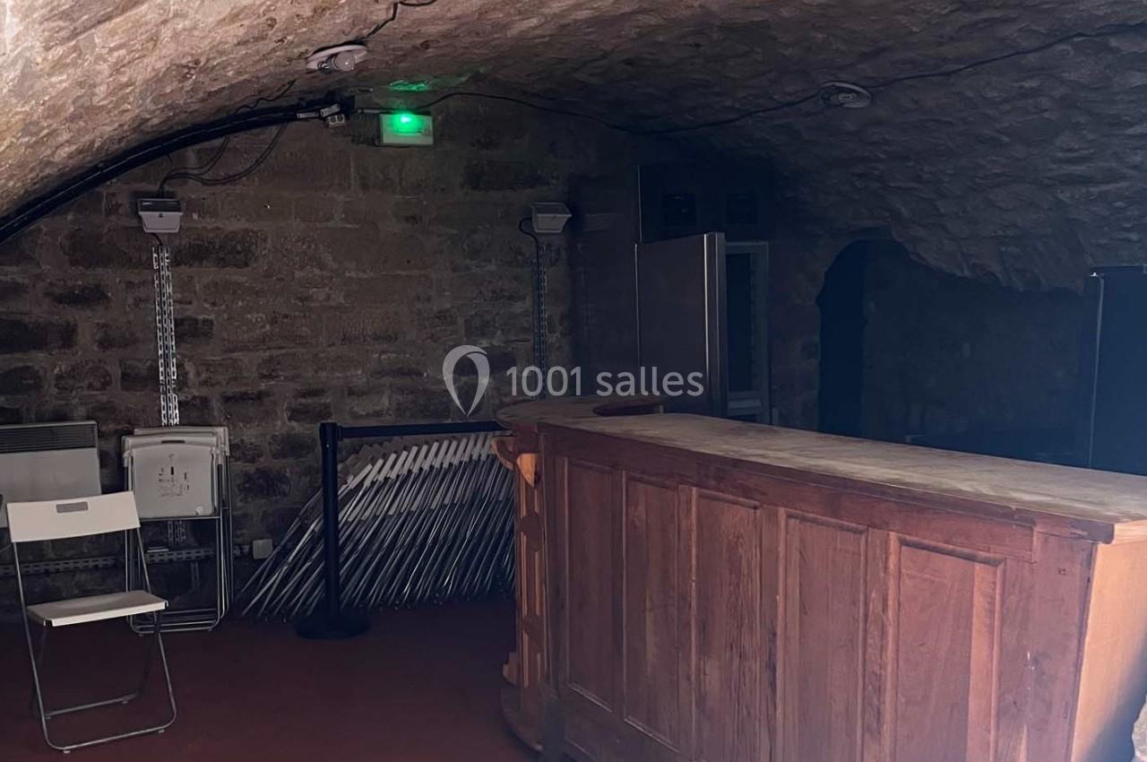 Un comptoir en bois dans une pièce voûtée en pierre avec des chaises pliantes empilées contre le mur.