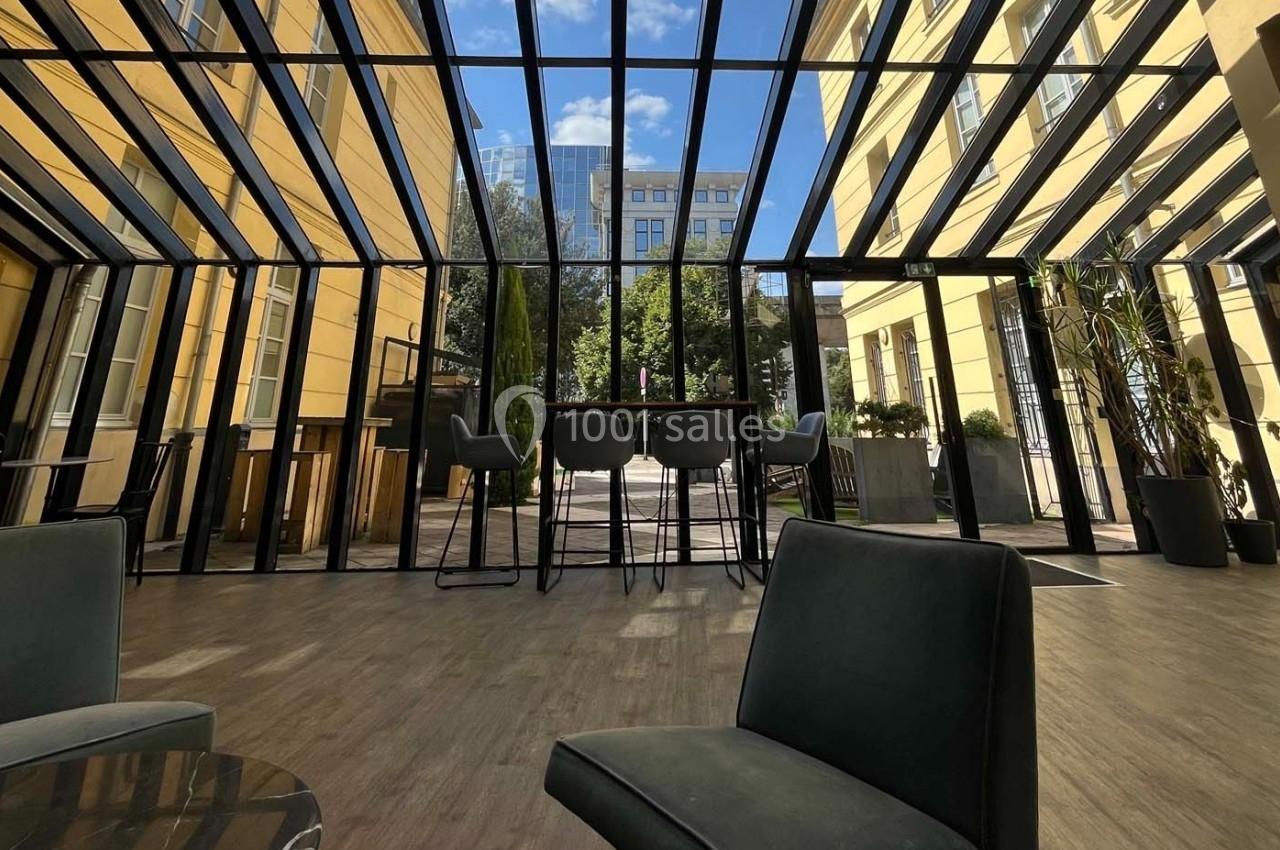 Espace intérieur lumineux avec verrière, mobilier moderne et vue sur une cour arborée par temps ensoleillé.