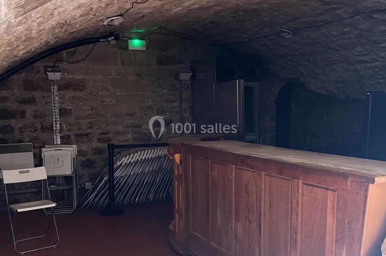 Cave voûtée en pierre avec un comptoir en bois, des chaises pliées et un éclairage tamisé.