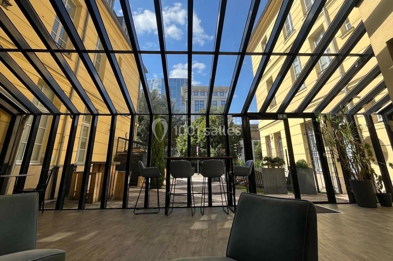 Espace intérieur lumineux avec verrière, mobilier moderne et vue sur une cour arborée sous un ciel bleu.