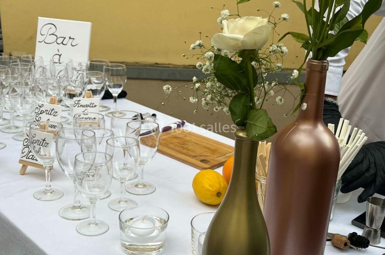 Table dressée avec des verres, bouteilles décoratives, une rose blanche et des citrons, devant un panneau ’Bar’.
