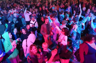 Homme en chemise noire, lunettes de soleil, fumant un cigare, tenant un verre devant un bar rempli de bouteilles.
