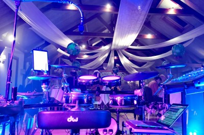 Homme en chemise noire, lunettes de soleil, fumant un cigare, tenant un verre devant un bar rempli de bouteilles.