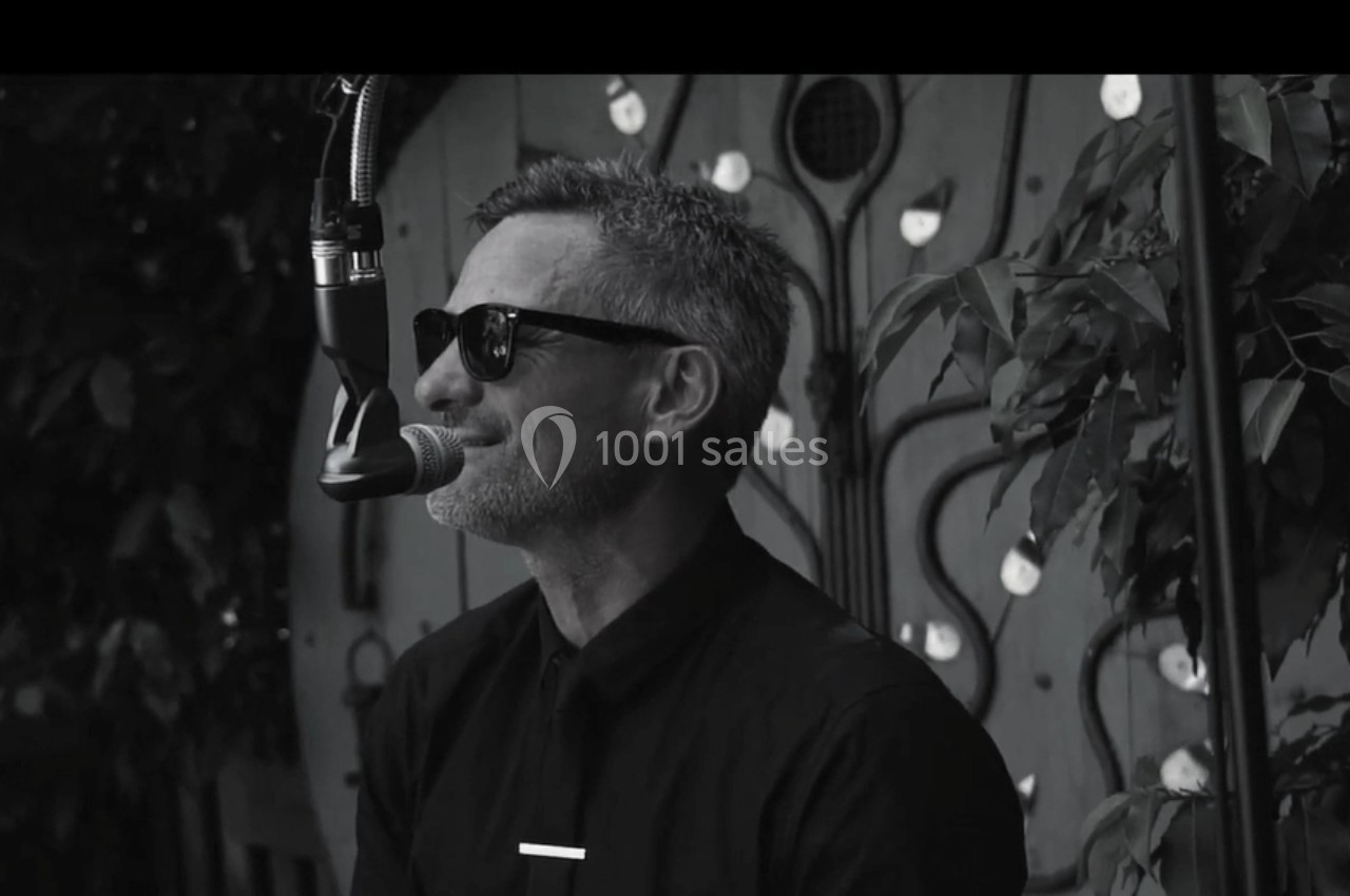 Homme portant des lunettes de soleil, chantant au microphone devant un décor végétal et artistique.