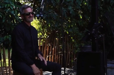 Homme en chemise noire, lunettes de soleil, fumant un cigare, tenant un verre devant un bar rempli de bouteilles.