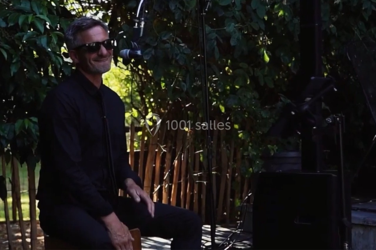 Un musicien souriant joue du cajón en extérieur, entouré de verdure et équipé d'un micro.