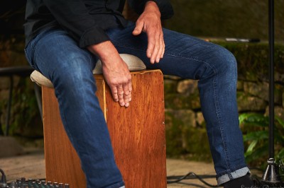 Homme en chemise noire, lunettes de soleil, fumant un cigare, tenant un verre devant un bar rempli de bouteilles.