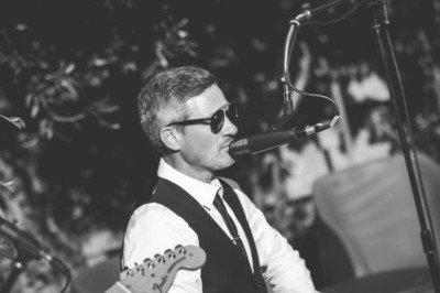 Homme en chemise noire, lunettes de soleil, fumant un cigare, tenant un verre devant un bar rempli de bouteilles.