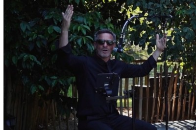 Homme en chemise noire, lunettes de soleil, fumant un cigare, tenant un verre devant un bar rempli de bouteilles.