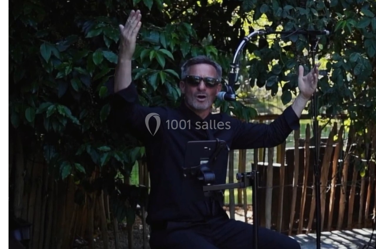 Un homme en lunettes de soleil chante ou parle au micro, bras levés, devant un fond de végétation et une clôture en bois.