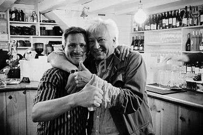 Deux hommes souriants s'enlacent dans un bar, entourés de bouteilles et de verres sur des étagères en bois.