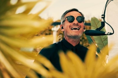 Homme souriant portant des lunettes de soleil, chantant au micro en extérieur, entouré de feuilles jaunes floues au premier…