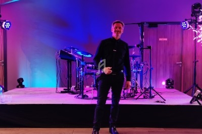 Un homme debout sur une scène éclairée en violet, entouré d'équipements de musique et de projecteurs.