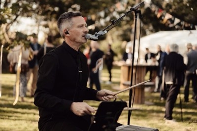 Un musicien joue de la batterie et chante au micro lors d'un événement en plein air avec des invités en arrière-plan.