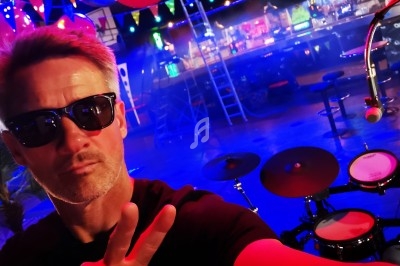 Homme portant des lunettes de soleil faisant un signe de paix devant une batterie dans une salle colorée et décorée.