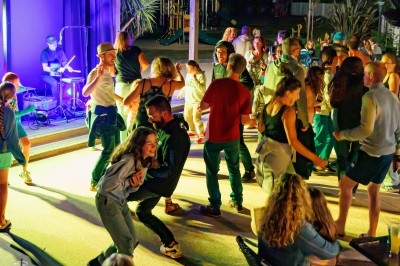 Groupe de personnes dansant et socialisant en soirée, éclairées par des lumières colorées près d'une scène avec un musicien.