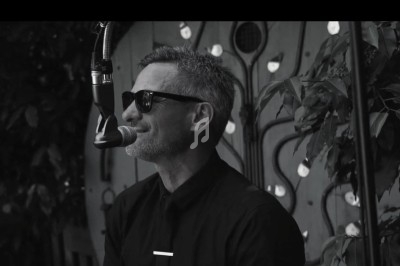 Homme portant des lunettes de soleil, chantant au microphone devant un décor végétal et artistique.