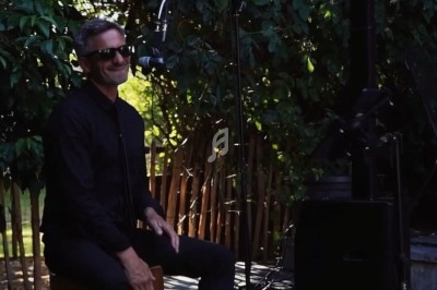 Un musicien souriant joue du cajón en extérieur, entouré de verdure et équipé d'un micro.