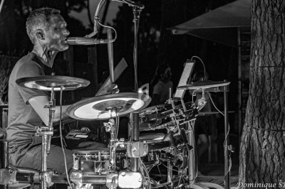 Un musicien joue de la batterie électronique et chante au micro lors d'une performance en extérieur, de nuit.