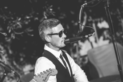 Un homme en costume et lunettes de soleil chante au micro lors d'une performance musicale en extérieur.