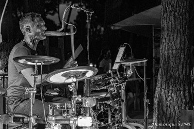 Homme en chemise noire, lunettes de soleil, fumant un cigare, tenant un verre devant un bar rempli de bouteilles.