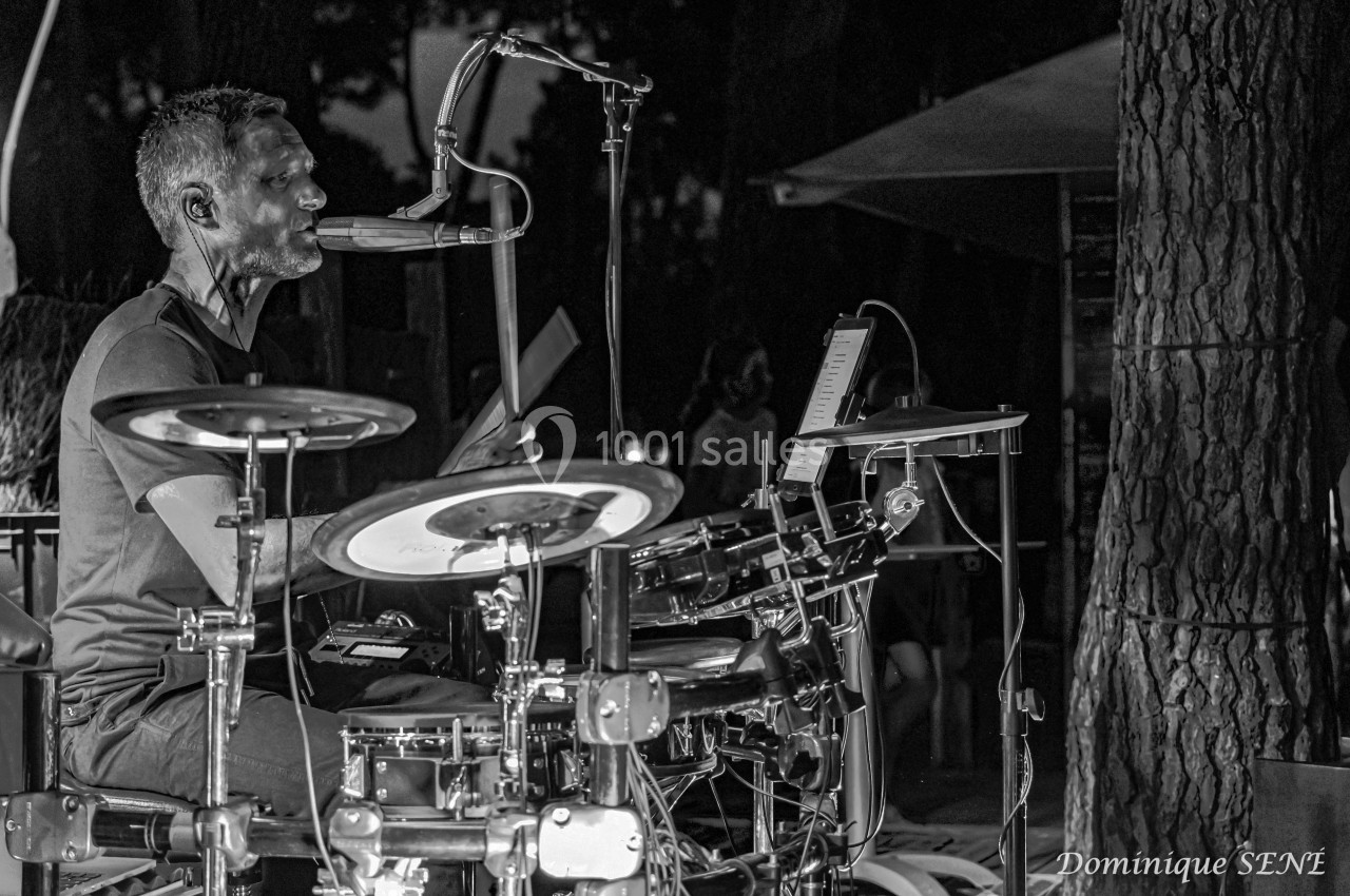 Un batteur joue sur une batterie électronique en extérieur, éclairé dans une ambiance nocturne.
