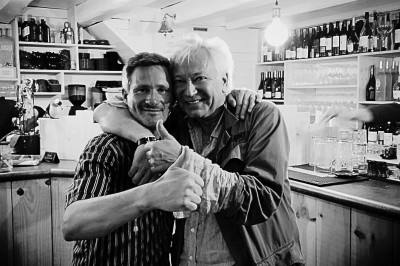 Homme en chemise noire, lunettes de soleil, fumant un cigare, tenant un verre devant un bar rempli de bouteilles.