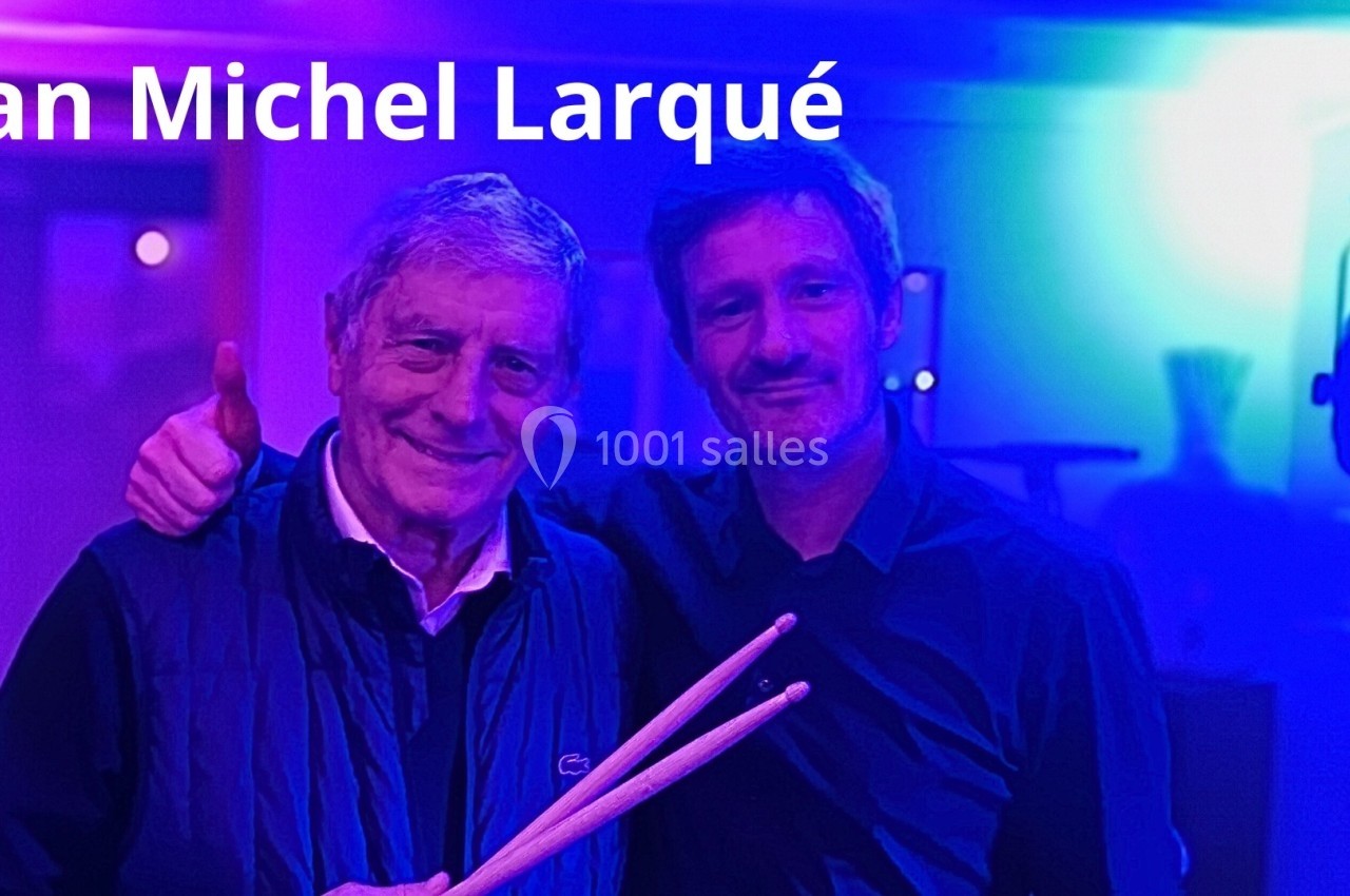 Deux hommes posent ensemble dans une ambiance lumineuse bleutée, l'un tenant des baguettes de batterie.