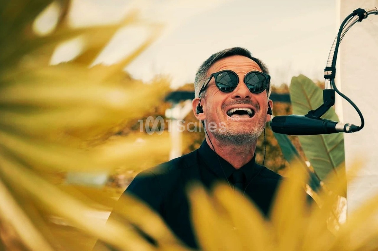 Un homme portant des lunettes de soleil chante au micro, entouré de feuilles floues au premier plan.