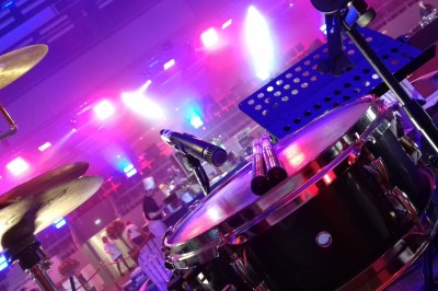 Homme en chemise noire, lunettes de soleil, fumant un cigare, tenant un verre devant un bar rempli de bouteilles.