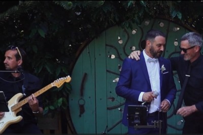 Homme en chemise noire, lunettes de soleil, fumant un cigare, tenant un verre devant un bar rempli de bouteilles.