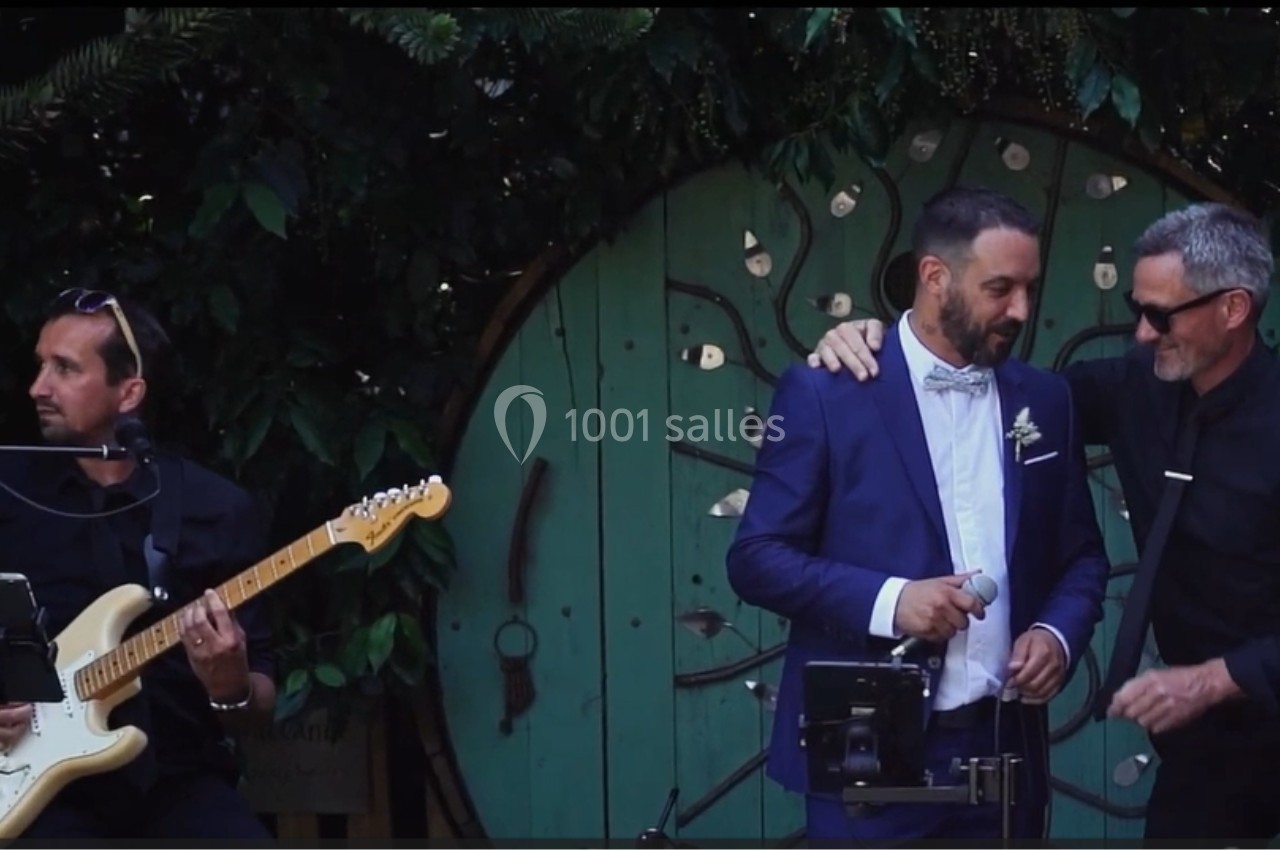 Un homme en costume bleu et un autre en noir se tiennent près d'un micro, avec un guitariste jouant à gauche.
