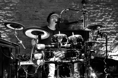 Homme en chemise noire, lunettes de soleil, fumant un cigare, tenant un verre devant un bar rempli de bouteilles.