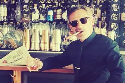 Homme en chemise noire, lunettes de soleil, fumant un cigare, tenant un verre devant un bar rempli de bouteilles.