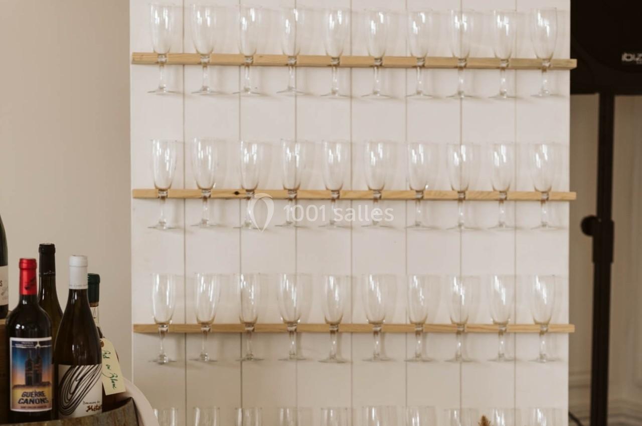 Des verres à vin vides alignés sur des étagères en bois fixées à un mur blanc, avec des bouteilles au premier plan.