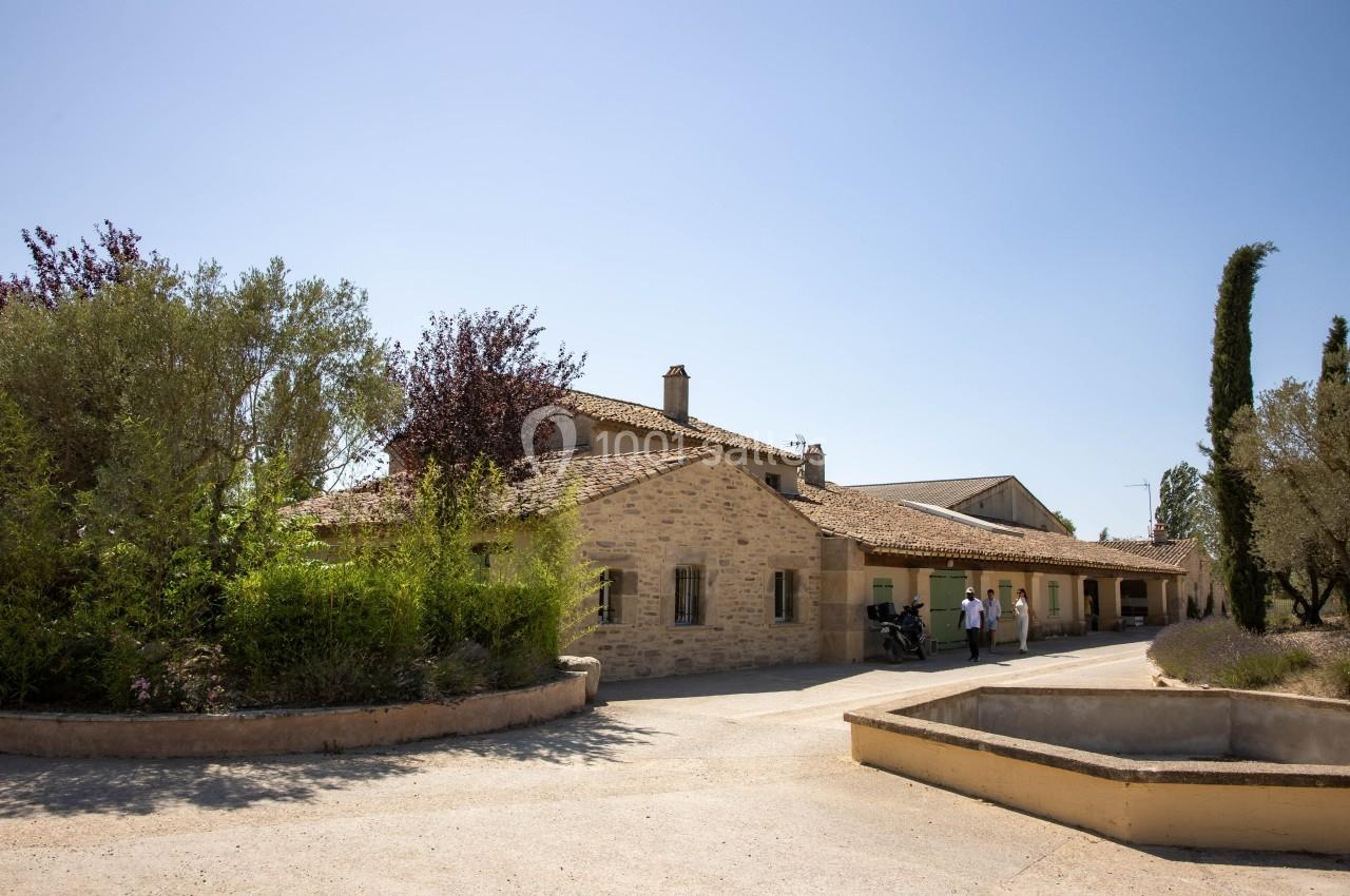 Bâtiment en pierre entouré de végétation méditerranéenne sous un ciel dégagé, avec quelques personnes à l'entrée.
