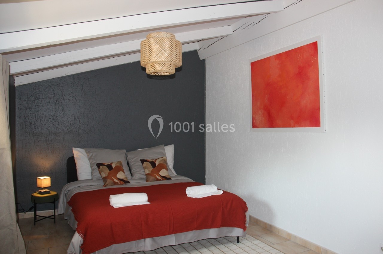 Chambre mansardée avec lit double, couvre-lit rouge, mur gris, tableau abstrait rouge et lampe suspendue en osier.